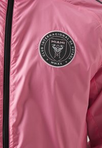 Giacca rosa chiaro con texture liscia, caratterizzata da un logo circolare nero con la scritta "MIAMI" e un'emblema. La giacca ha una cerniera frontale.