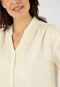 Blusa crema con ricami floreali, colletto classico e chiusura con bottoni. Tessuto morbido con una leggera texture e un design leggero.