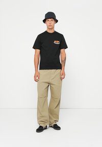 Sort T-shirt med frontgrafik, beige brede bukser, sorte sko og en marineblå bøttehat. Ensfarvet baggrund; ingen synlige mønstre.