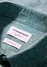Col de chemise en tissu texturé vert avec des étiquettes blanches indiquant Lindbergh relaxed fit et lin en coton.