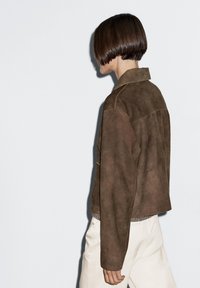 Hnědá semišová bunda s límcem a zkráceným střihem. Výrazné šití a měkká textura doplňují světle zbarvený top a kalhoty.