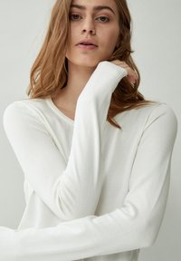 Giovane donna con capelli castano chiaro che indossa una maglia bianca a maniche lunghe, posando con una mano vicino al viso su uno sfondo neutro.