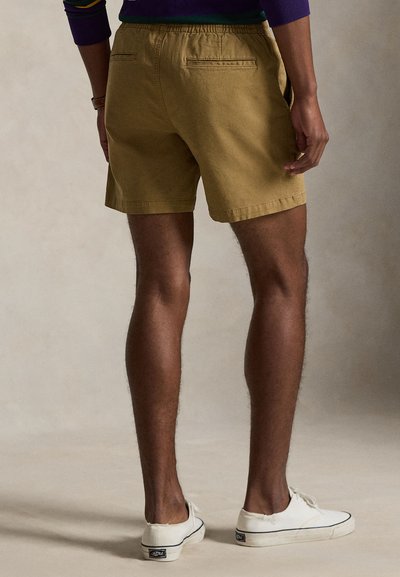 Háttal álló férfi, világosbarna rövidnadrágot, fehér tornacipőt és lila hosszú ujjú felsőt visel, egyszerű háttér előtt.