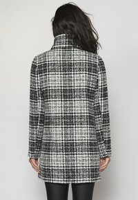Dlouhý černobílý károvaný kabát s vysokým límcem, vyrobený z texturované látky s přímým střihem, sahající do poloviny stehen.