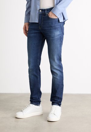 Osoba nosząca niebieskie jeansy typu slim-fit, białe trampki, niebieską koszulę, z jedną ręką w kieszeni, stojąca na szarej betonowej podłodze na białym tle.