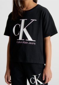 Černé bavlněné tričko s volným střihem, které má velké bílé logo "CK" a menší text "Calvin Klein Jeans" v růžové barvě pod logem.