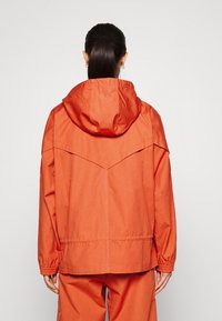 Orange lättviktsjacka med huva, som har elastiska ärmslut och en lös passform. Texturerad tyg med en sydd detalj över ryggen.