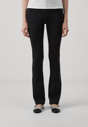 Black slim-fit pants with a smooth texture and subtle front seams, paired with black and silver flats, against a neutral background.