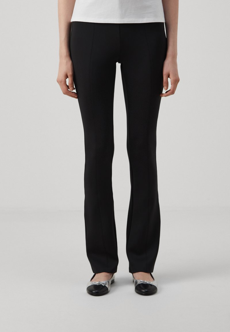 Črne slim-fit hlače z gladko teksturo in subtilnimi sprednjimi šivi, v kombinaciji s črnimi in srebrnimi balerinkami, na nevtralni ozadju.