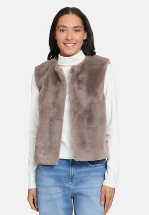 Femme souriante portant un gilet en fausse fourrure taupe par-dessus un col roulé blanc à côtes et un jean bleu clair, se tenant contre un fond uni clair.
