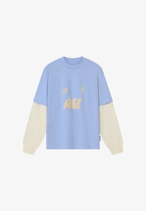 Garām piedurknēm krekls ar gaiši zilu korpusu un krēmveida piedurknēm, uz krūtīm un kreisās piedurknes tekstā ir "ACI" logotips un mazi dzelteni dizaini.