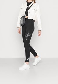 Zwarte legging met "AIR" in het wit gedrukt, gecombineerd met een cropped witte jas en witte sneakers. Textuur schoudertas.