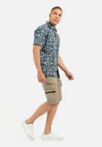 Korte mouwen blauwe bloemenoverhemd, kaki cargoshorts met ritsen, en witte sneakers. Heeft patronen van bladeren en vogels.