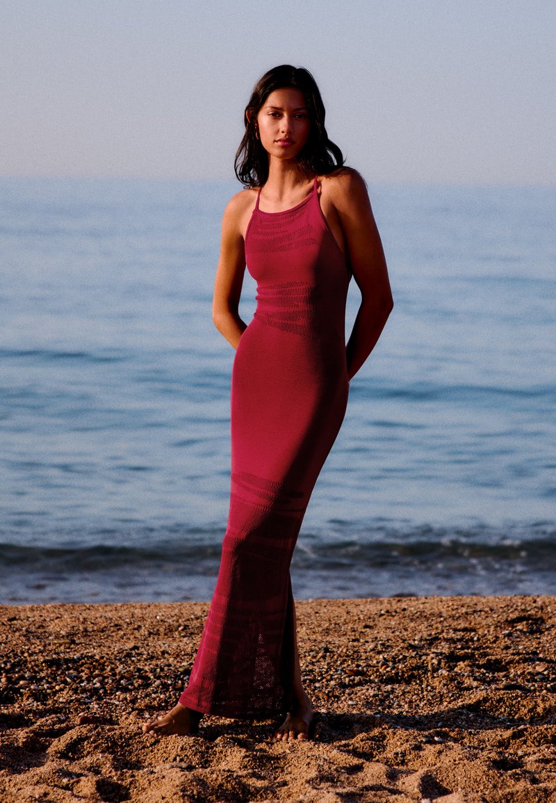 Femme en robe rouge ajustée sans manches, debout pieds nus sur une plage de sable avec une mer calme en arrière-plan.