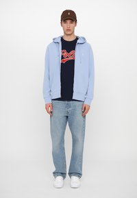 Sweatshirt azul claro com fecho, t-shirt gráfica azul-marinho com o logótipo "Polo", jeans de denim claros e ténis brancos. Boné castanho com detalhe do logótipo.