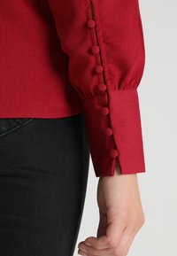 Rød bluse med lange ærmer, der har et tekstureret stof, pufærmer og en knaplukning med runde stofbetrukne knapper ved manchetterne.