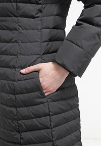 Black padded coat featuring horizontal quilted sections, a side pocket, and a smooth finish. Sleeve with a distinct cuff detail.
