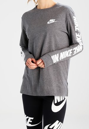 Kobieta nosząca szary, długi rękaw Nike z białymi logo na klatce piersiowej i rękawach, połączona z czarnymi legginsami Nike z dużymi białymi logo.