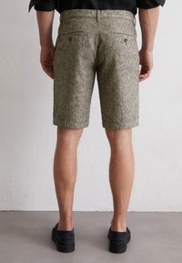 Grün-beige gestreifte Shorts aus strukturiertem Stoff, mit zwei Gesäßtaschen und einer taillierten Passform, die knapp über dem Knie endet. Schwarze Schuhe.