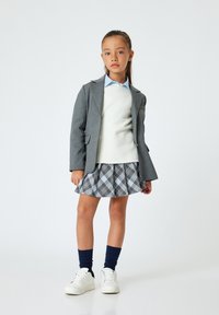Blazer grigio sopra un maglione bianco e una camicia azzurro chiaro, abbinato a una gonna a quadri con tonalità blu e grigie, scarpe da ginnastica bianche e calze blu navy.