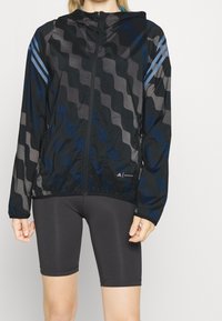 Giacca nera con un motivo ondulato in tonalità di grigio e blu. Presenta un cappuccio, zip completa e due tasche laterali. Il materiale sembra leggero.