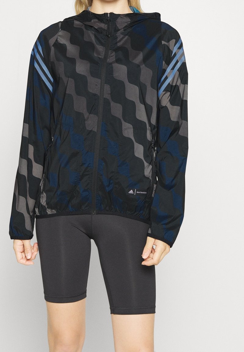 Giacca nera con un motivo ondulato in tonalità di grigio e blu. Presenta un cappuccio, zip completa e due tasche laterali. Il materiale sembra leggero.