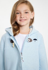 Ljusblå zip-up hoodie med en mjuk innerfoder, med broderade detaljer av ett öga och ett paraply. Dragkedja med en dekorativ dragdel.