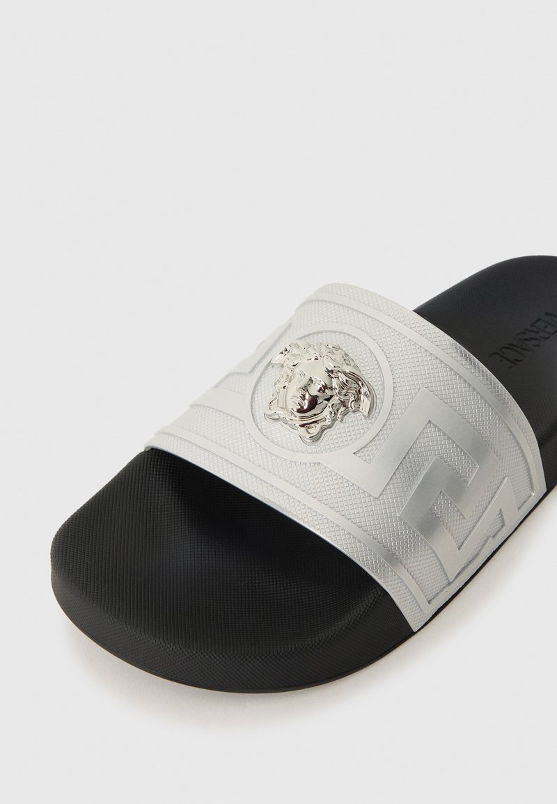 Silverfärgad slidesandal med ett strukturerat rem med en Medusa-emblem och en svart gummisula med rundad kant. Enkel geometrisk struktur.