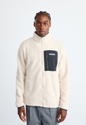 Fleece jacket in cream with a black zippered chest pocket. Features a high collar and side pockets. Soft texture, relaxed fit.