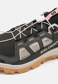 Melna un pelēka sieta sporta kurpe ar baltu auklu sistēmu, rozā akcentiem un izturīgu gumijas zoli ar saķeres elementiem. Redzama Salomon zīmola logo.