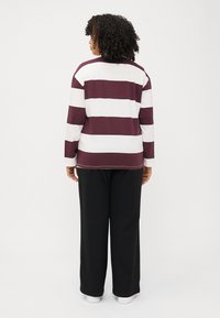 Une personne aux cheveux bouclés se tient de dos, portant une chemise à rayures maroon et blanche à manches longues et un pantalon noir, se tenant devant un fond blanc.