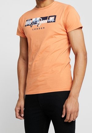 Mężczyzna ubrany w dopasowany brzoskwiniowy t-shirt z krótkim rękawem z logo „Pepe Jeans London” oraz czarne spodnie na jednolitym tle.