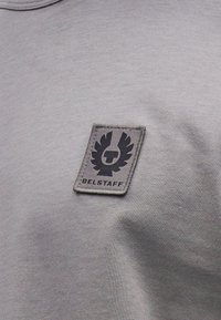 Pilkos medvilninės marškinėliai, ant krūtinės srityje siuvinėtas juodas odinis lopas su sparnuotu logotipu ir „BELSTAFF“ tekstu.
