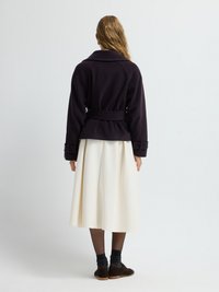 Giacca viola scuro con vita cinturata, dotata di un ampio colletto e maniche con risvolti, abbinata a una gonna bianca e lunga fino al ginocchio e scarpe marroni.