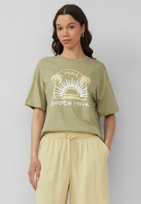 Oliwkowa koszulka z krótkim rękawem, z grafiką słońca i palm, z napisem "Złota Godzina" w białym kolorze. Noszona z jasnopomarańczowymi spodniami.