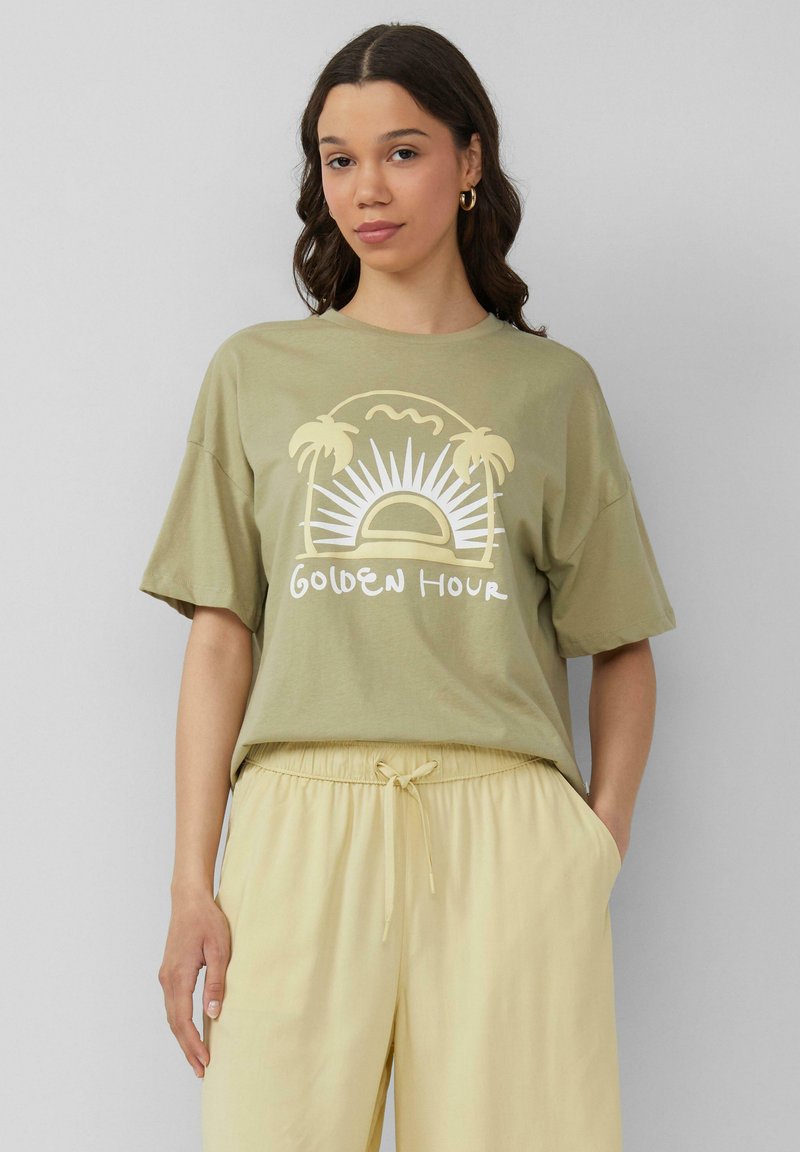 Oliwkowa koszulka z krótkim rękawem, z grafiką słońca i palm, z napisem "Złota Godzina" w białym kolorze. Noszona z jasnopomarańczowymi spodniami.