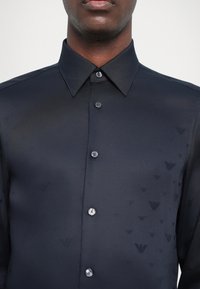 Camicia blu marina con un sottile motivo a triangoli, maniche lunghe e colletto a punta. Presenta una chiusura con bottoni abbinati.