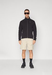 Musta fleece-takki vetoketjulla edessä, vaaleat shortsit, mustat sukat ja kengät. Malli, jolla on aurinkolasit, seisoo yksinkertaisella taustalla.