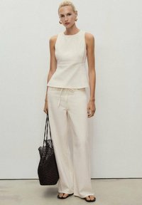 Kremowa bluzka peplum w parze z luźnymi, kremowymi spodniami o szerokich nogawkach. Modelka trzyma brązową torebkę plecioną; cały look dopełniają proste czarne klapki.