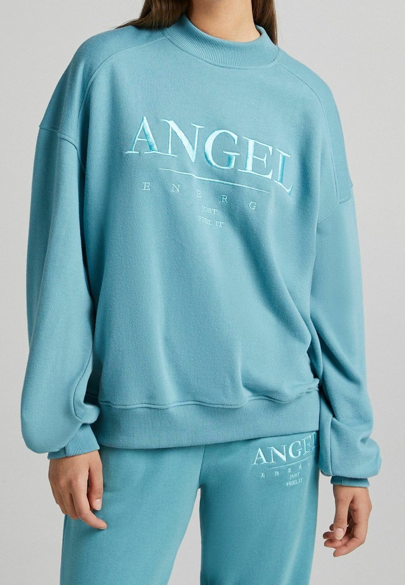 Turkos sweatshirt med präglad "ANGEL"-logotyp, ribbad halsringning och ärmslut, avslappnad passform, matchas med en par joggers. Slät textur.