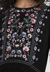 Top de tela negra con bordado floral intrincado en rosa, azul y blanco. El diseño incluye patrones florales y un adorno decorativo.