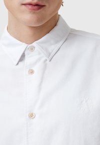 Hvid skjorte med knapper har en klassisk krave og diskret logo-broderi. Den er lavet af glat bomuld, har en struktureret overflade og naturlige knapper.