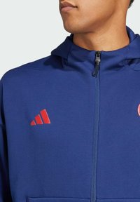 Sudadera con capucha de color azul marino con cremallera completa, presenta un logotipo rojo de tres rayas en el lado izquierdo del pecho y una tela texturizada. Puños y dobladillo elásticos.