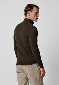 Ciemnozielony, lekki, dopasowany sweter z wysokim kołnierzem i ściągaczem u dołu, noszony z beżowymi spodniami, pokazany od tyłu.