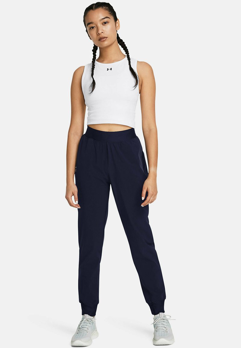 Weißes, cropped Tanktop mit schwarzem Logo, kombiniert mit marineblauen Jogginghosen mit elastischem Bund und schmal zulaufenden Bündchen. Graue Sportschuhe.