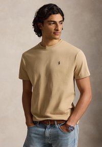 Young man with curly dark hair wears a beige short-sleeve shirt and blue jeans, hands in pockets, looking slightly to the side.