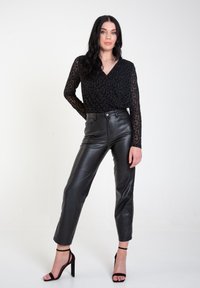 Black, long-sleeve top with leopard print, paired with high-waisted black leather trousers. Model wears black stiletto heels, posing with hands on hips.
