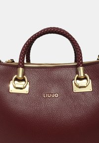 Borsa in pelle bordeaux con una finitura testurizzata, manico intrecciato, dettagli in metallo dorato e logo del marchio sulla parte anteriore. Chiusura con zip.