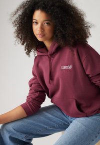 Burgundyröd huvtröja med frontficka och broderad logotyp. Matchad med ljusblå, löst sittande jeans.