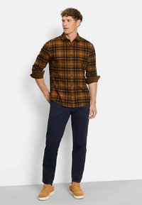 Braun-schwarz kariertes Hemd mit langen Ärmeln und Knopfleiste, kombiniert mit dunkeln marineblauen Hosen und hellbraunen Sneakers. Schlichter Hintergrund.